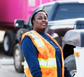 A mature African-American woman wearing a safety vest working at a commercial dock. She is a truck driver standing beside her semi-truck. She is looking at the camera, smiling.